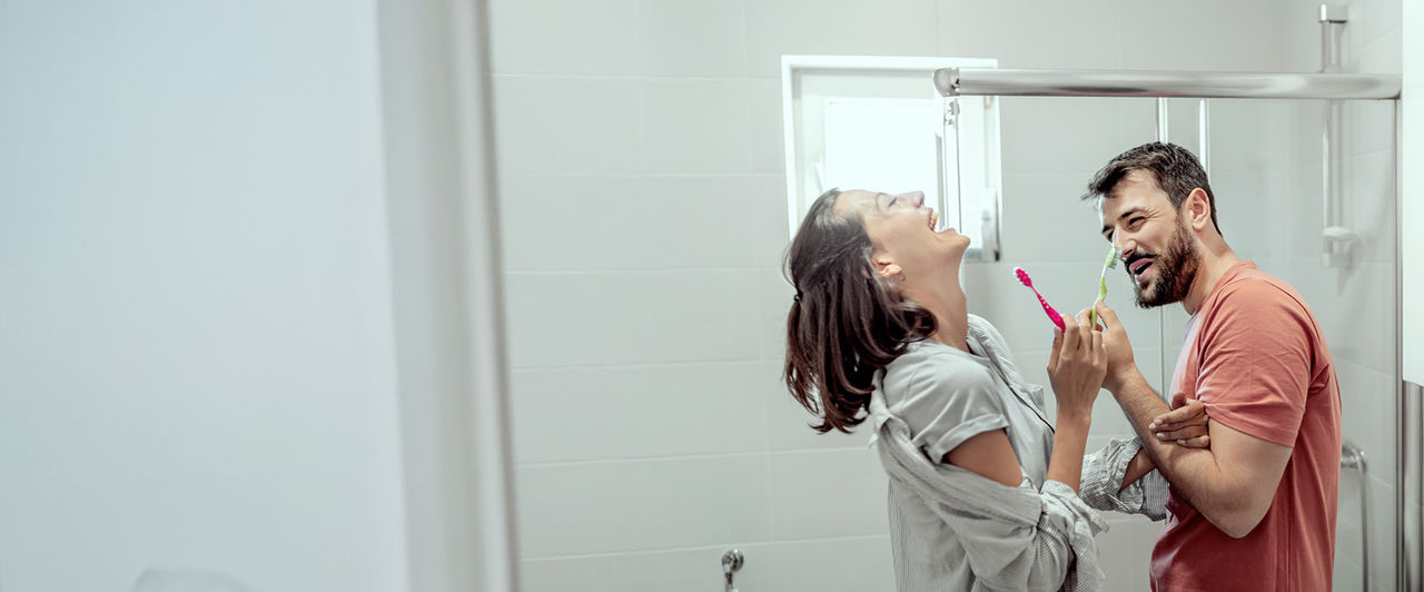 Pareja riendo en el baño mientras sostienen cepillos de dientes coloridos, promoviendo la higiene bucal y el bienestar diario en el hogar.