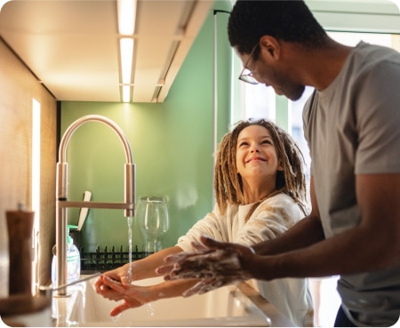 Un hombre frente al fregadero de una cocina enseñándole a un niño cómo lavarse las manos correctamente como parte de una rutina saludable de cuidado personal.