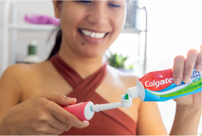 Una mujer sonríe mientras pone pasta de dientes Colgate en un cepillo de dientes en un cuarto de baño.